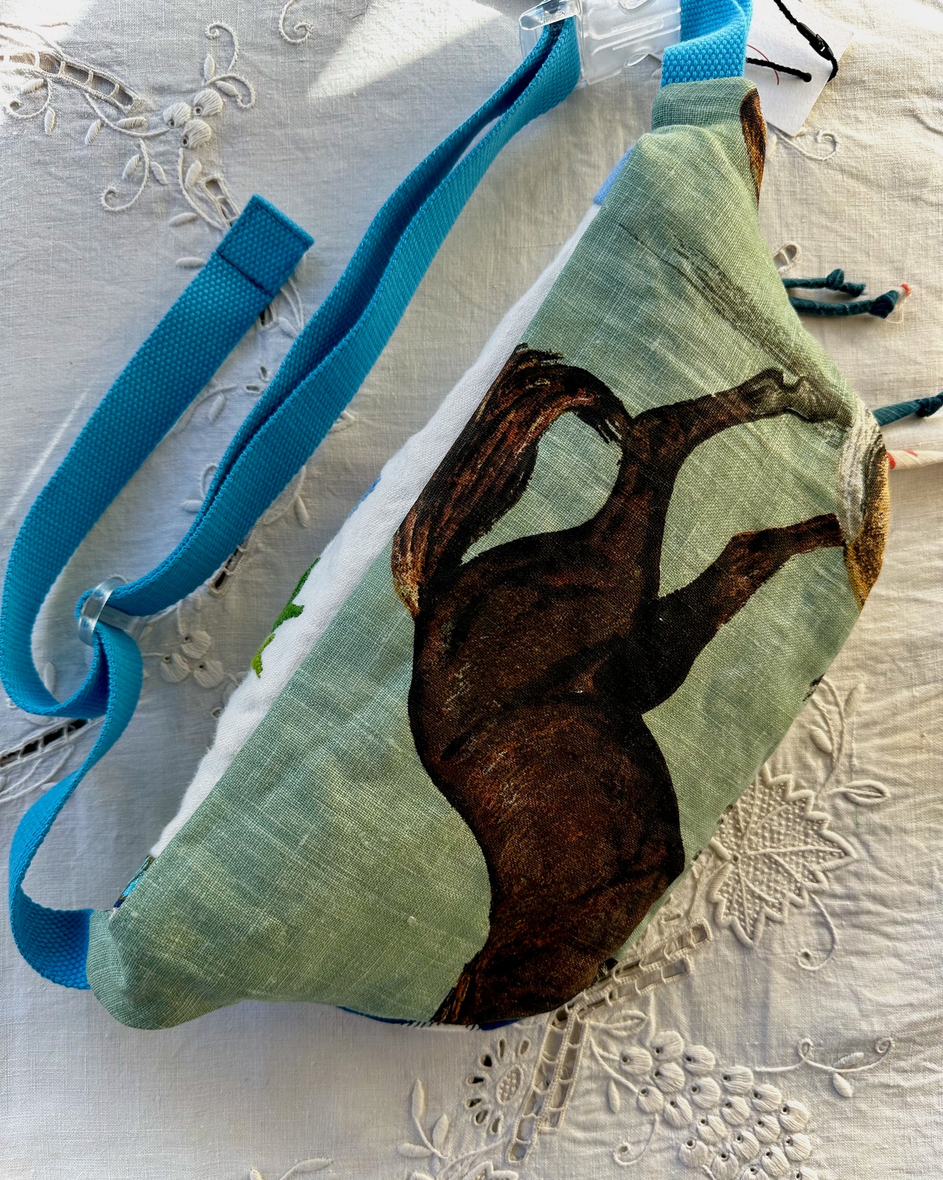 A fabric crossbody bag with a light green background and a dark brown horse illustration, featuring a bright blue strap, laid flat on a white embroidered fabric surface.