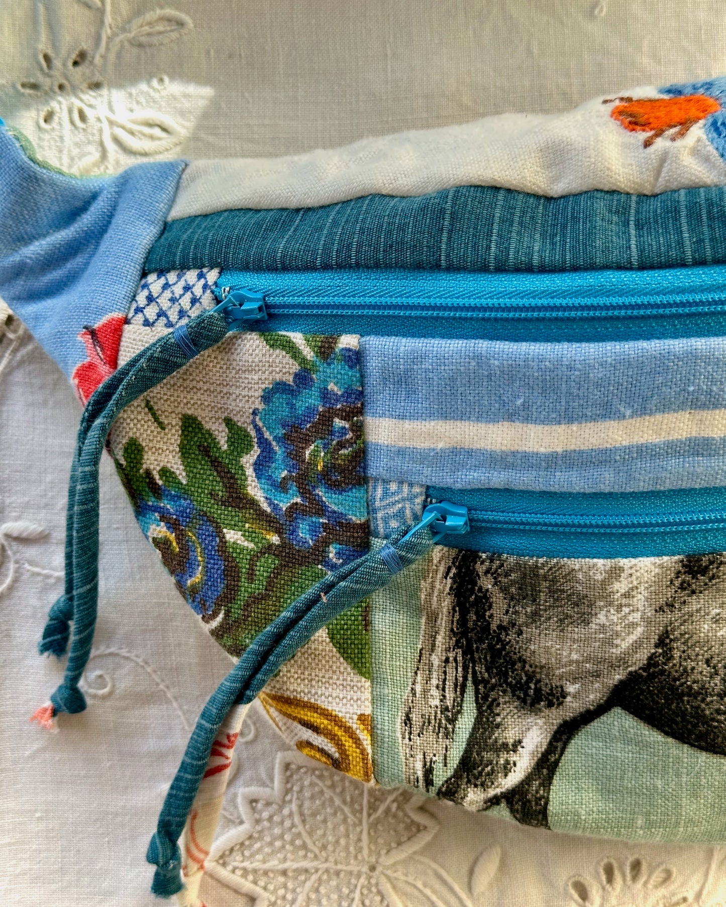 A close-up of a handmade patchwork fanny pack with a blue zipper, featuring various fabric panels including blue stripes, a floral print, and a horse design, resting on a white embroidered fabric surface.