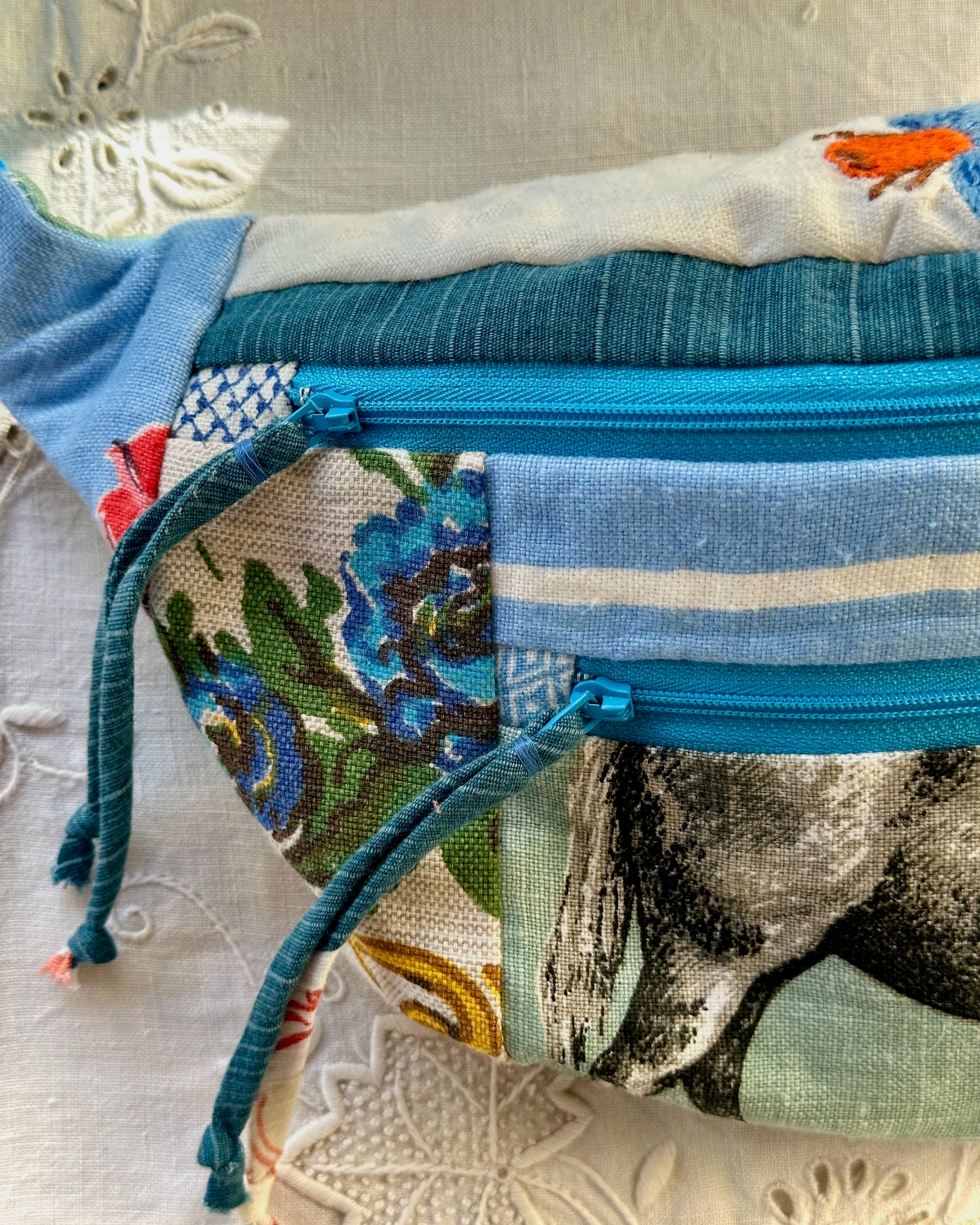 A close-up of a handmade patchwork fanny pack with a blue zipper, featuring various fabric panels including blue stripes, a floral print, and a horse design, resting on a white embroidered fabric surface.