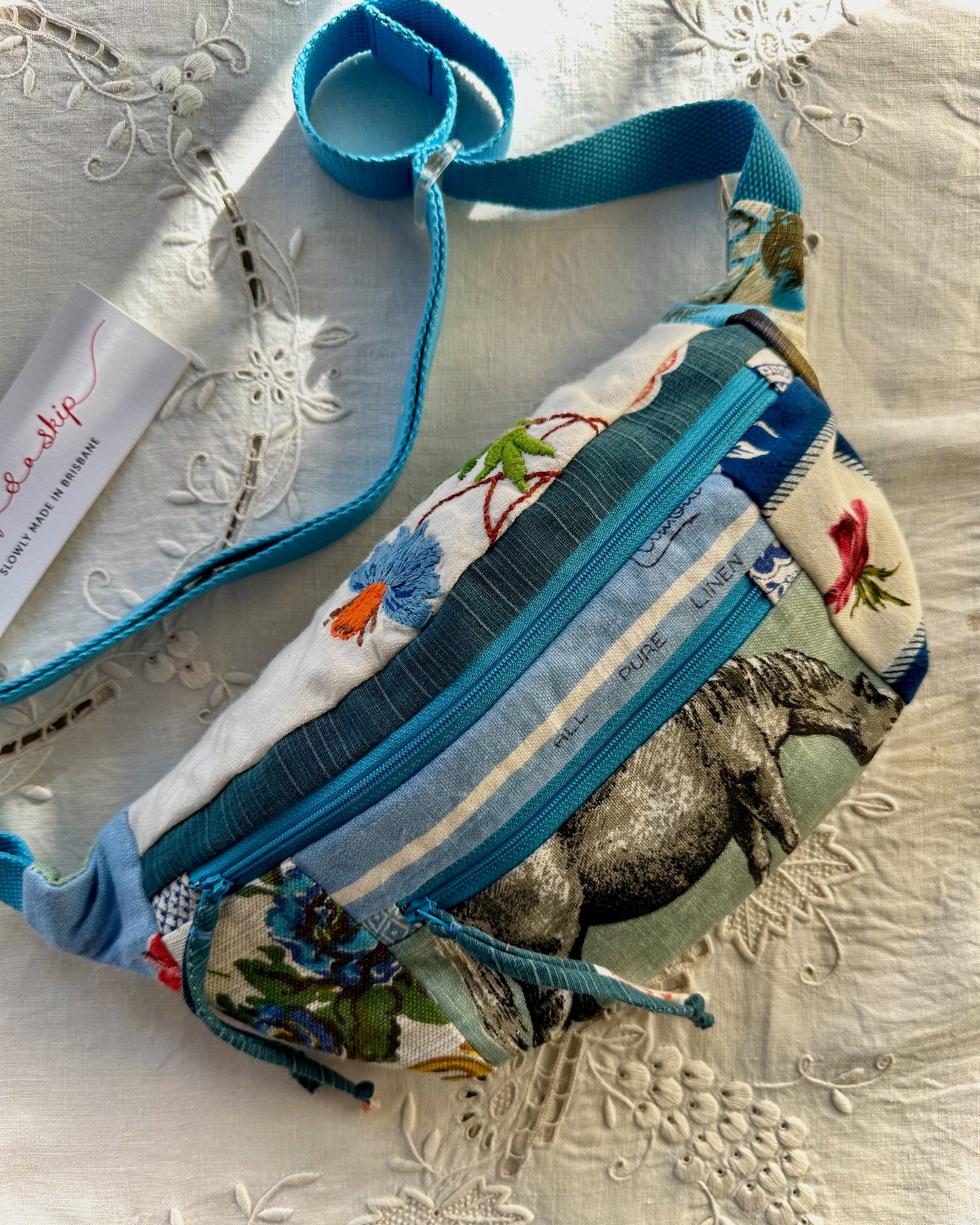 A fabric crossbody bag with a colorful animal and floral print, featuring bright blue trim and a matching blue strap, laid flat on a white embroidered fabric surface.