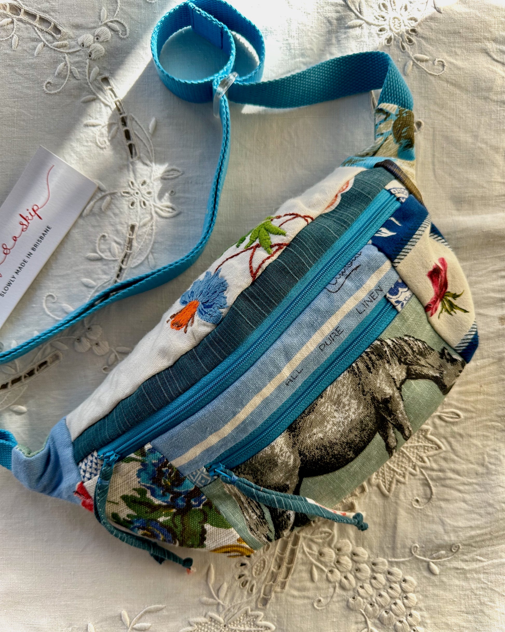 A fabric crossbody bag with a colorful animal and floral print, featuring bright blue trim and a matching blue strap, laid flat on a white embroidered fabric surface.