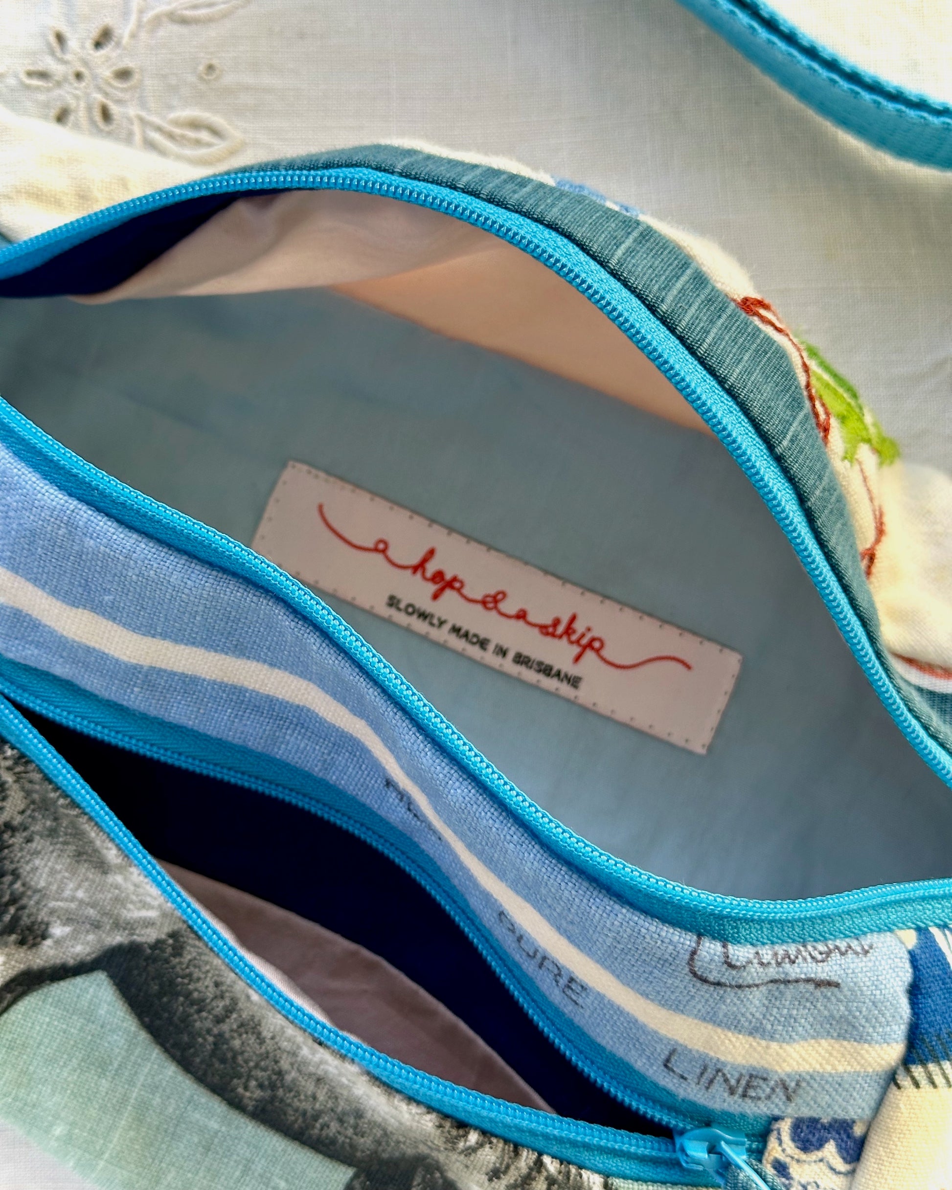 Close-up of an open zippered bumbag showing the interior. The exterior fabric has a blue and white patterned print, a bright blue zipper, and a visible sewn-in label reading 'a hop and a skip' with 'SLOWLY MADE IN BRISBANE' underneath.