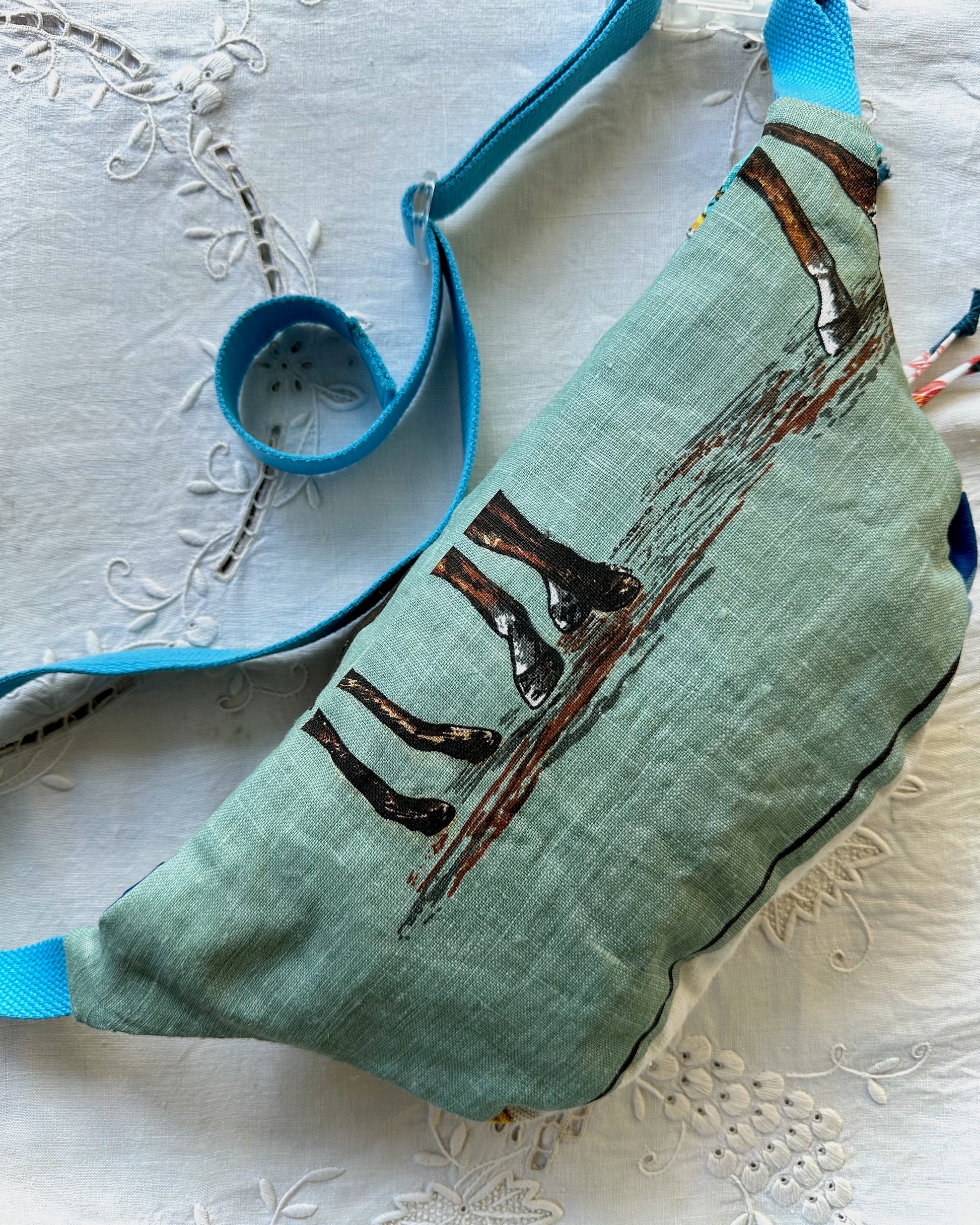 A mint green fabric crossbody bag with a printed design of horse hooves on a wooden path, featuring a bright blue strap, laid flat on a white embroidered fabric background.