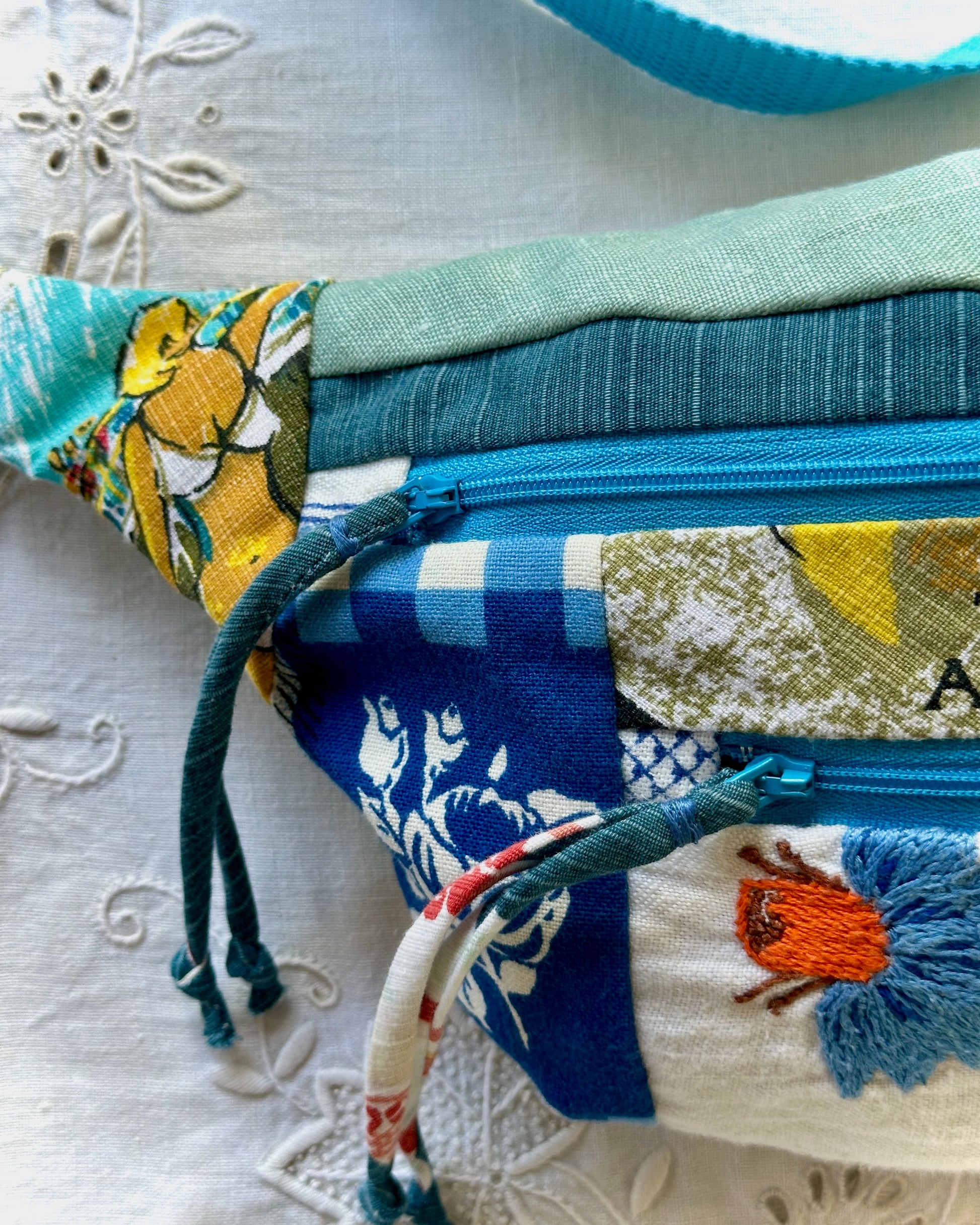 A close-up of a handmade patchwork fanny pack with a colorful floral and geometric print, featuring a blue zipper, dark green drawstring, and a light blue strap, laid on a white embroidered fabric surface.