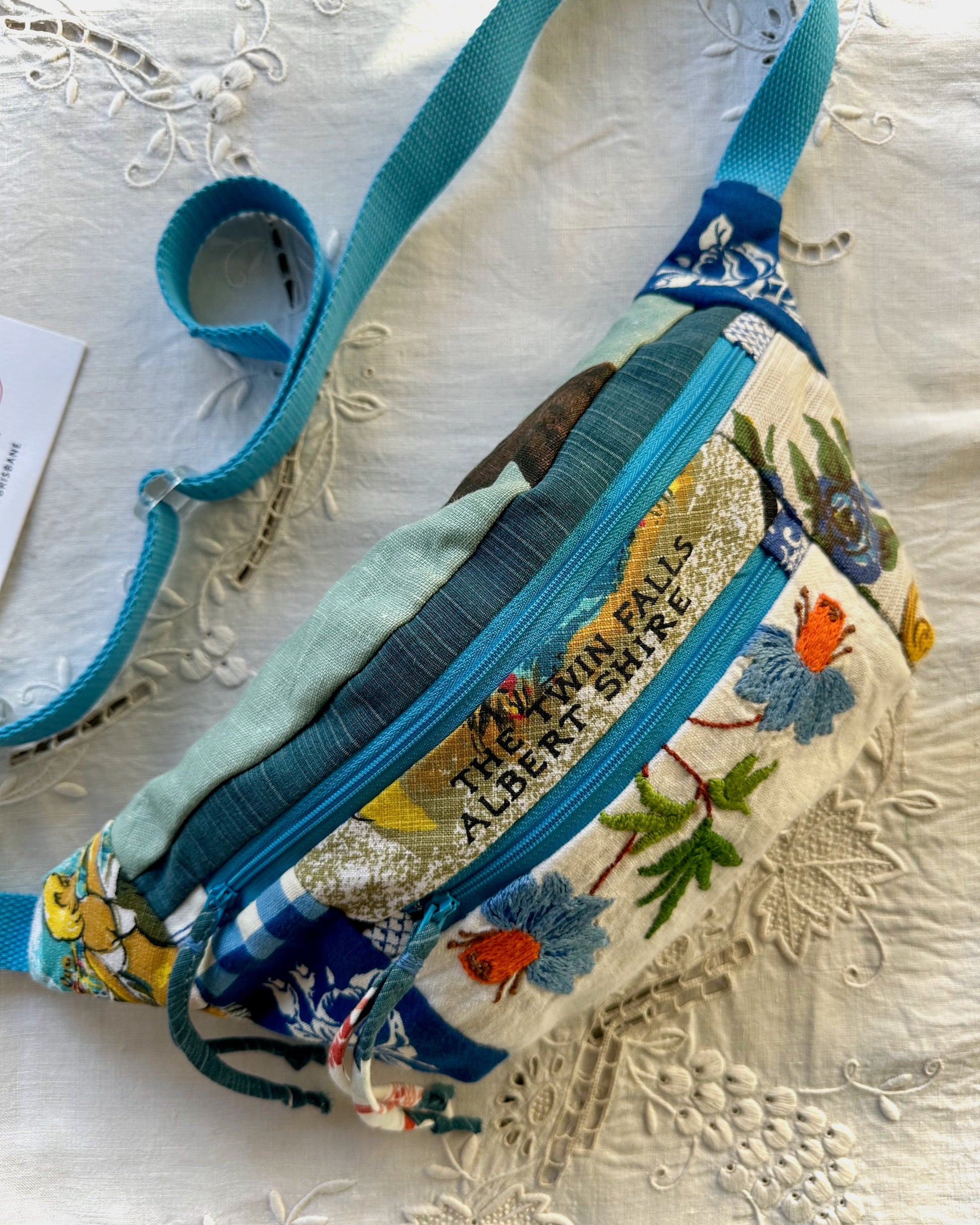 A handmade fabric bum bag with a turquoise strap and multiple patterned fabric panels, including floral embroidery and a gold glitter accent, resting on a white embroidered lace surface.