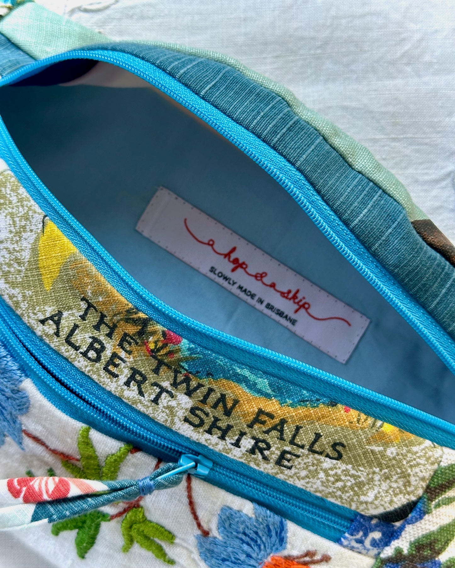 Close-up of a fabric zippered pouch or bag interior with a turquoise zipper, light blue lining, a white sewn label reading 'a hop and a skip', and a printed exterior section that says 'THE TWIN FALLS ALBERT SHIRE'.