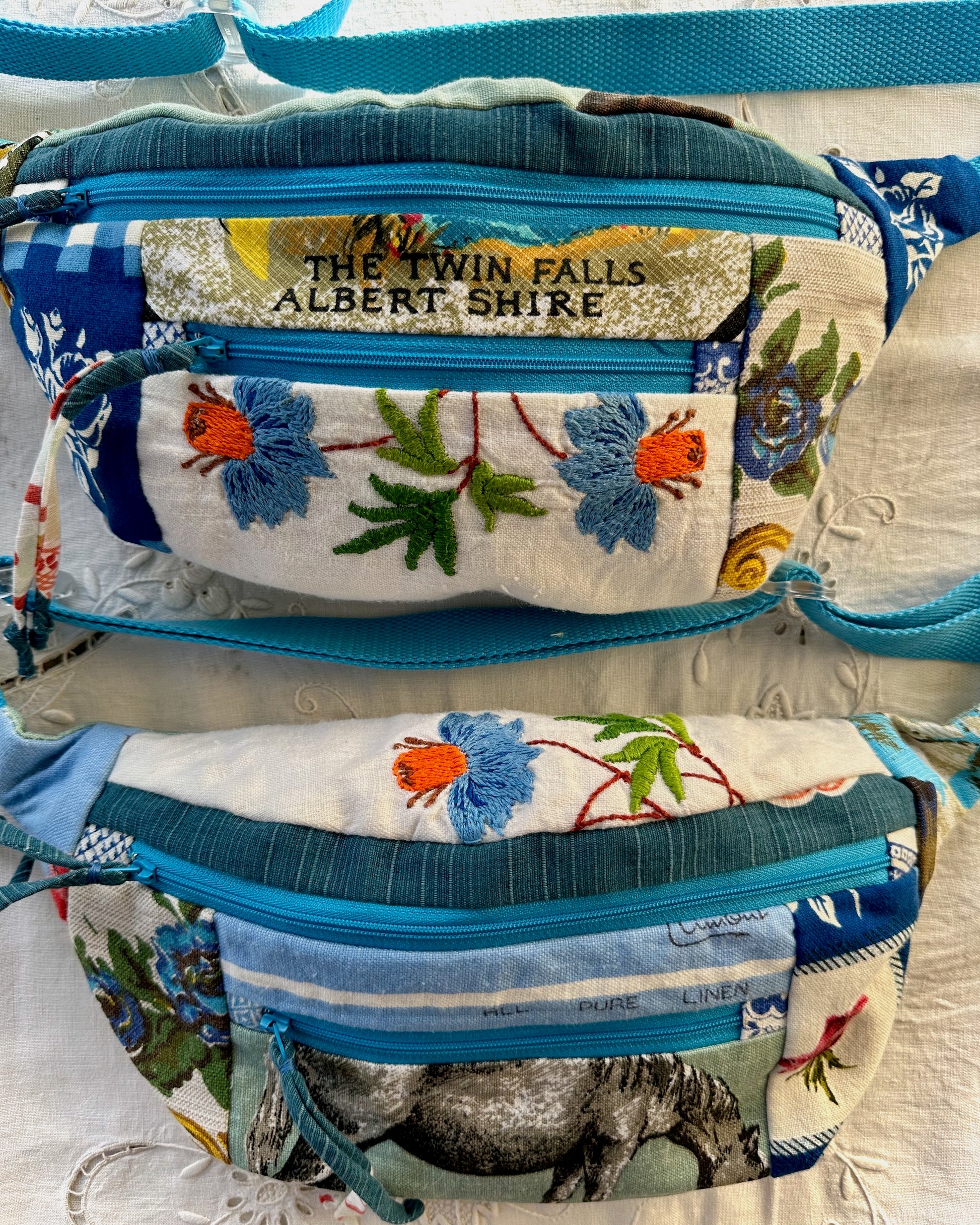 Two handmade fabric waist bags with blue zippers and colorful floral embroidery, displayed on a white textured fabric background. The top bag features a gold glitter banner reading 'THE TWIN FALLS ALBERT SHIRE'.