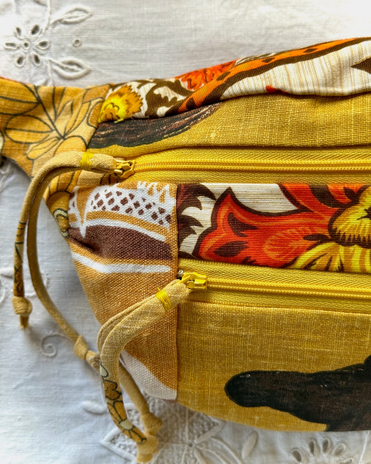 A close-up of a fabric waist bag with a multicolor patchwork print featuring mustard yellow, orange, brown, and cream floral and geometric patterns, with a yellow zipper and drawstring.