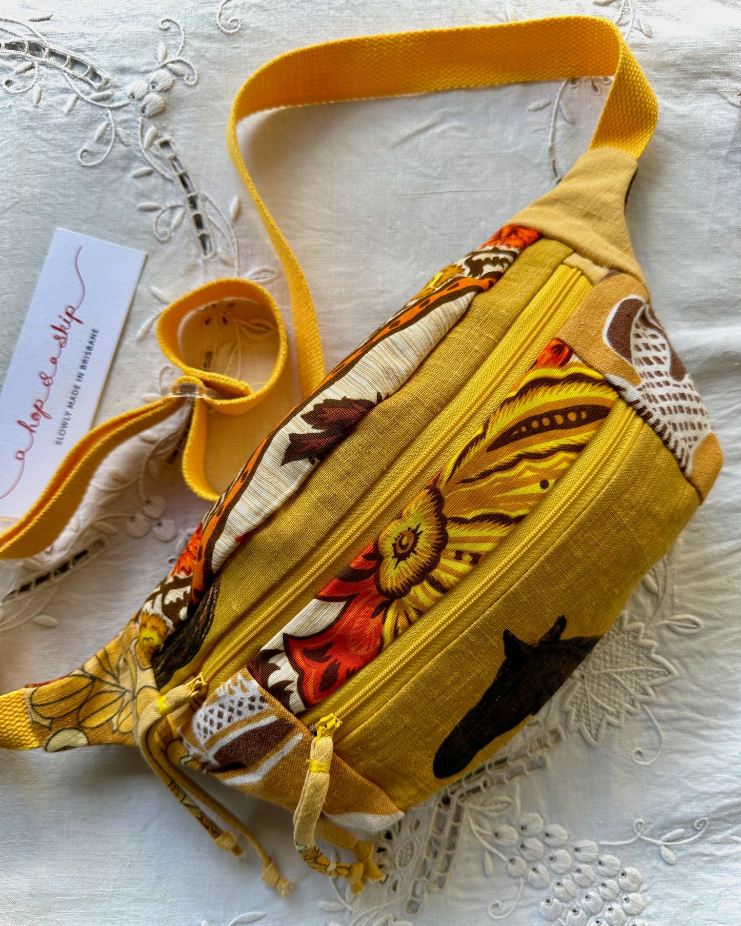 A yellow fabric crossbody bag with a floral and abstract print, featuring a wide yellow strap, a zippered front pocket, and drawstring details, displayed on a white embroidered fabric surface with a brand tag.