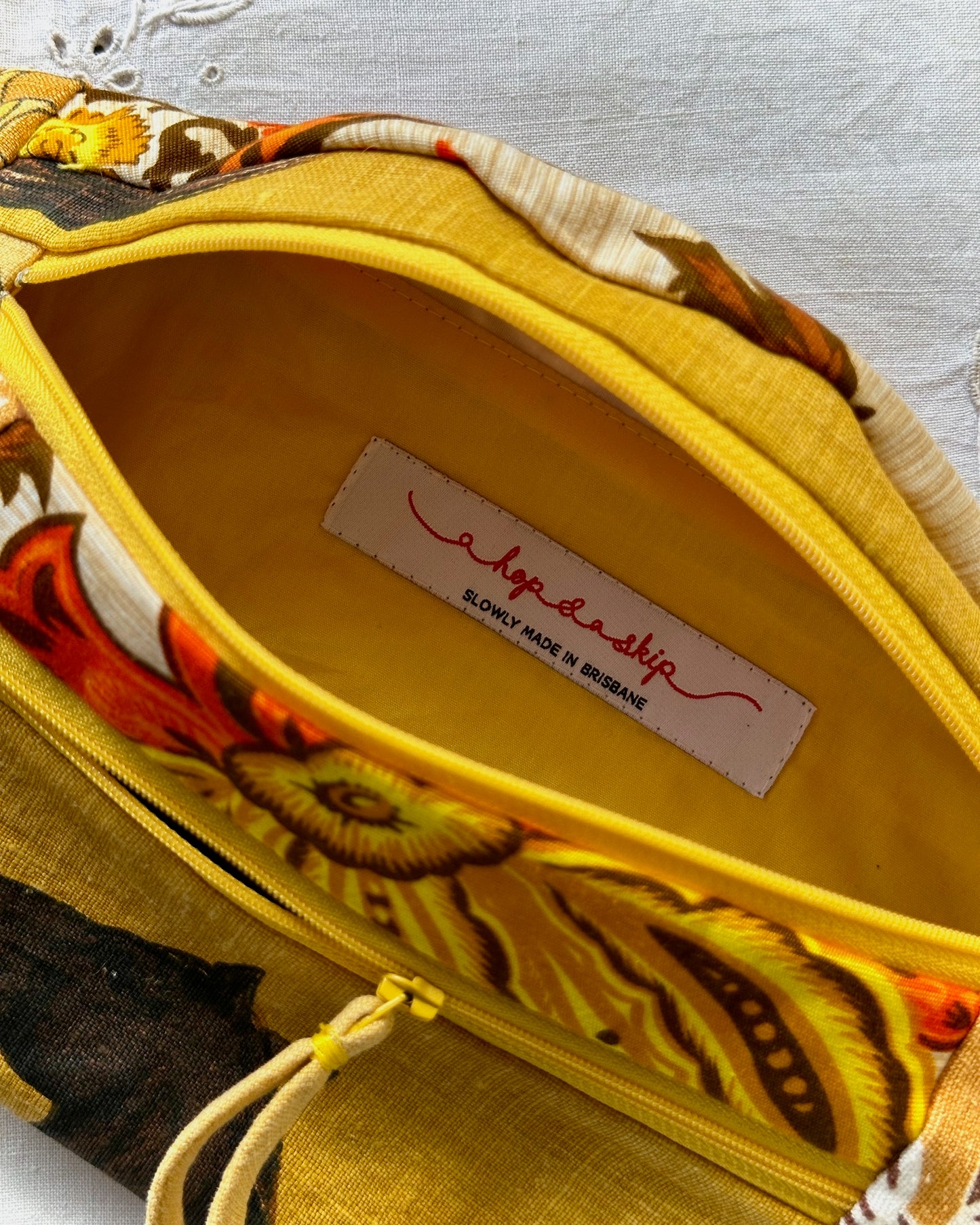 Close-up of a fabric bumbag interior with a bright yellow lining and a floral print exterior in yellow, orange, brown, and cream. A sewn-in label reads 'a hop and a skip' and 'SLOWLY MADE IN BRISBANE'.