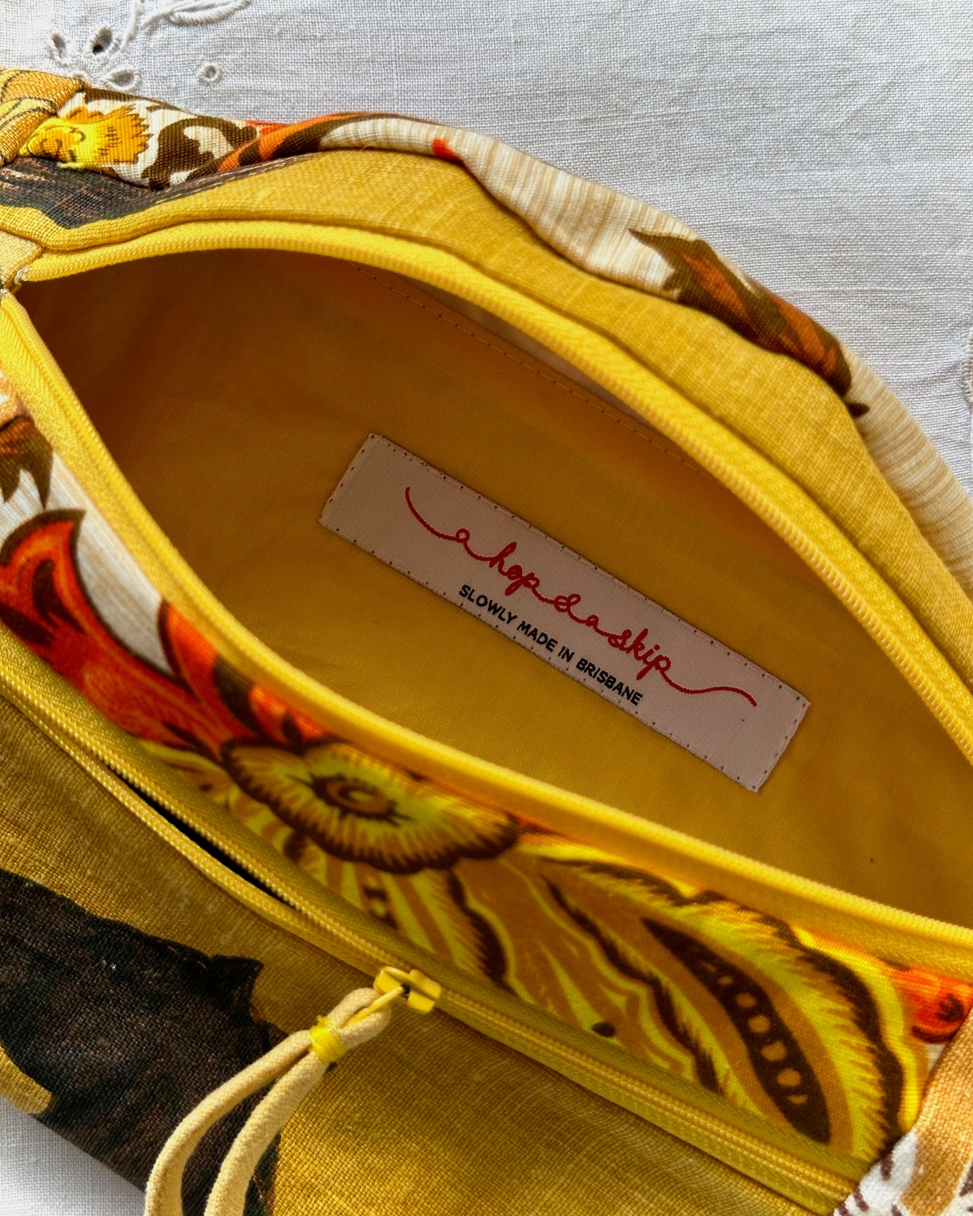 Close-up of a fabric bumbag interior with a bright yellow lining and a floral print exterior in yellow, orange, brown, and cream. A sewn-in label reads 'a hop and a skip' and 'SLOWLY MADE IN BRISBANE'.