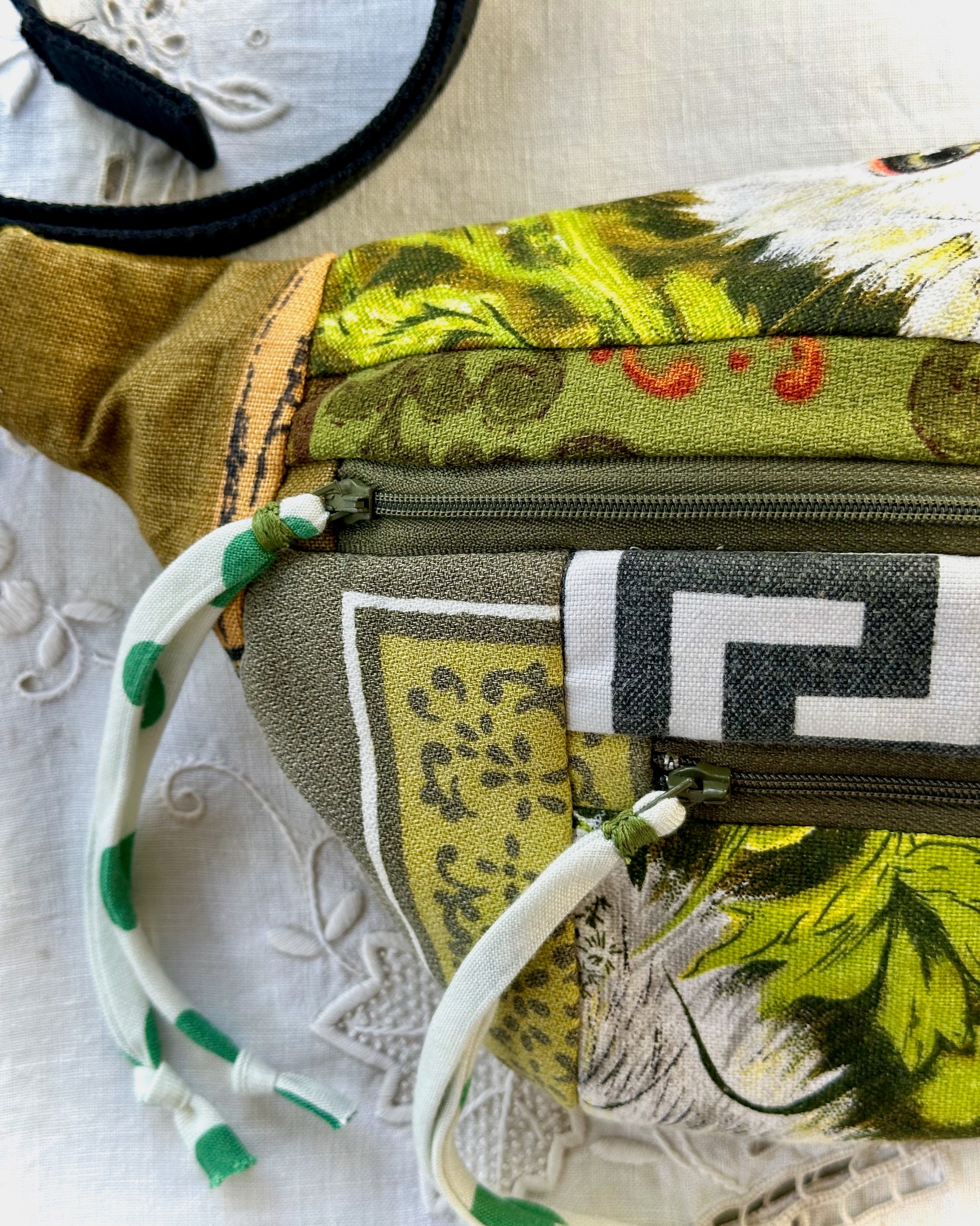 A close-up of a fabric bag or pouch with mixed prints, including olive green, yellow-green, black, white, and a Greek key pattern, on a white lace background.