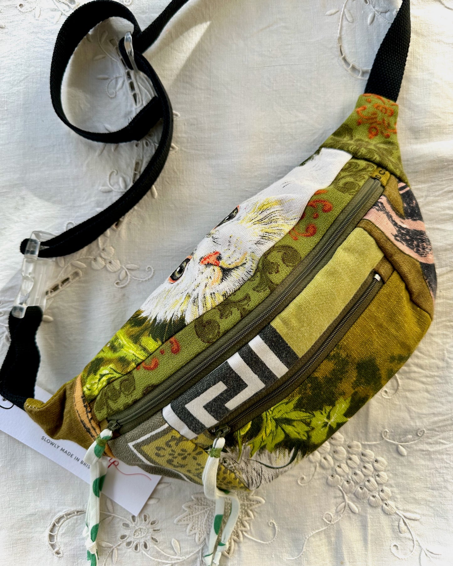 A fabric fanny pack with a multicolor print featuring a white cat, olive green patterns, and a black-and-white geometric stripe, shown on a white embroidered fabric background with its black strap and paper tag visible.