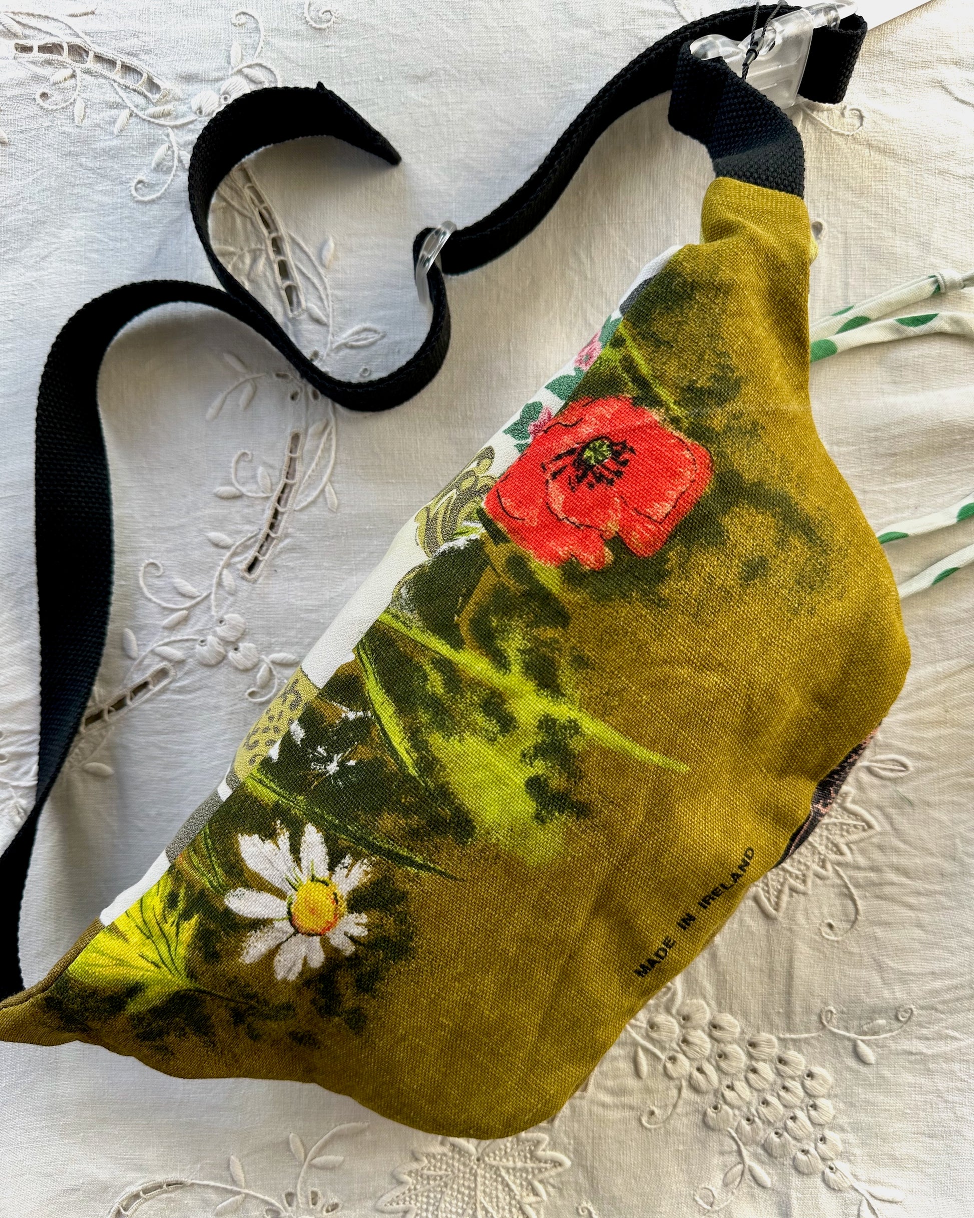 A fabric fanny pack or belt bag with a black adjustable strap, featuring a vintage-style botanical print in olive green, red poppy, and white daisy colors, laid flat on a white embroidered lace fabric background.