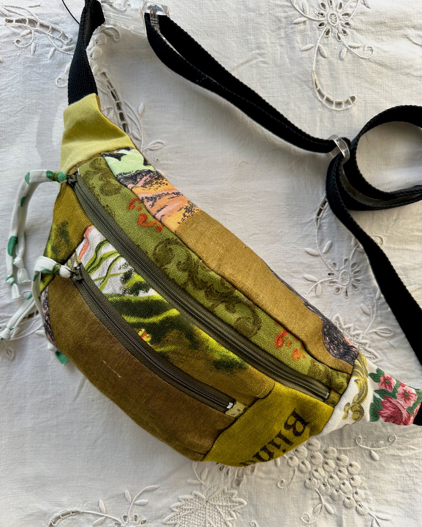 A handmade fabric fanny pack with a mixed print design in olive green, mustard yellow, and floral patterns, featuring a black adjustable strap, resting on a white embroidered lace fabric.