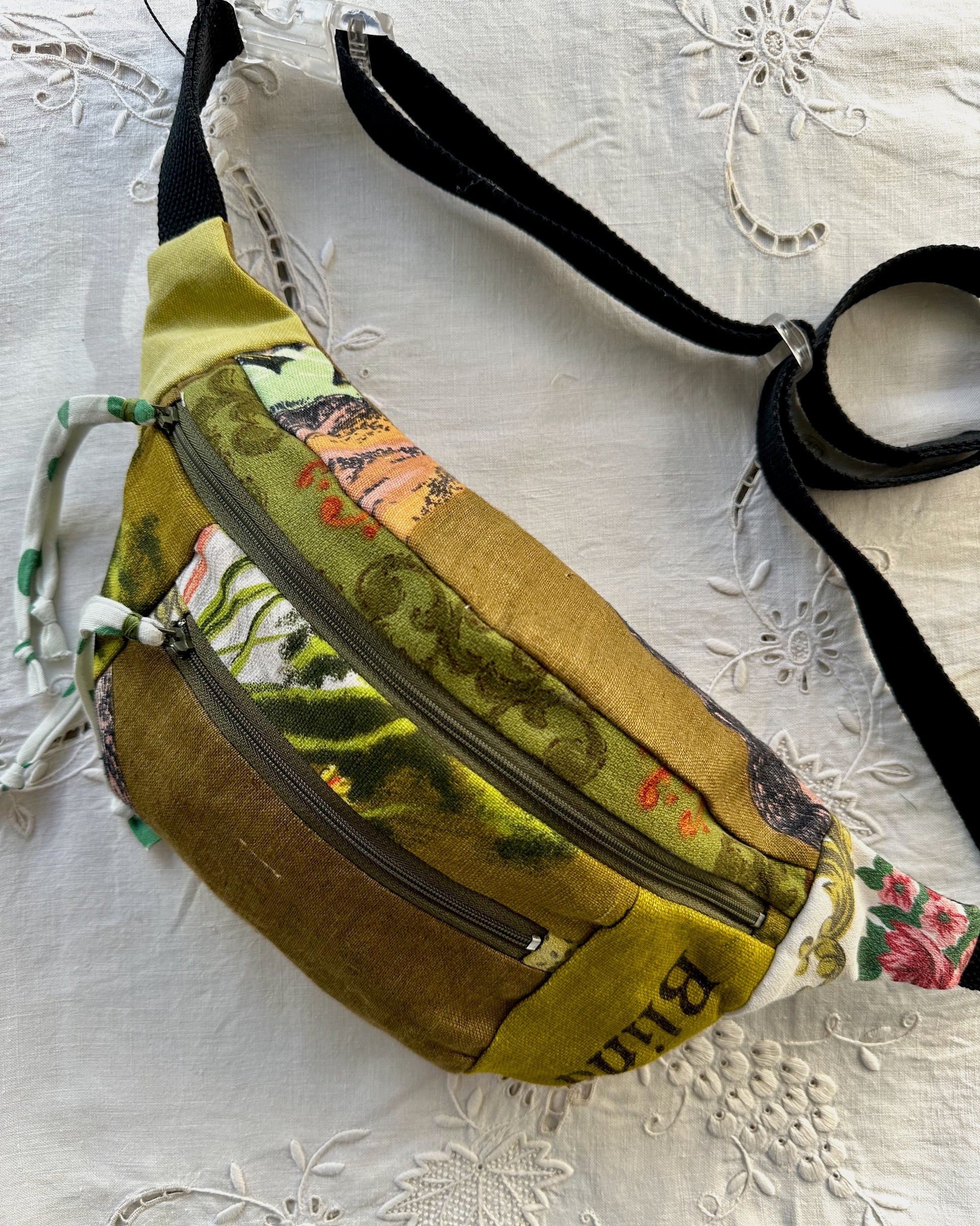 A handmade fabric fanny pack with a mixed print design in olive green, mustard yellow, and floral patterns, featuring a black adjustable strap, resting on a white embroidered lace fabric.