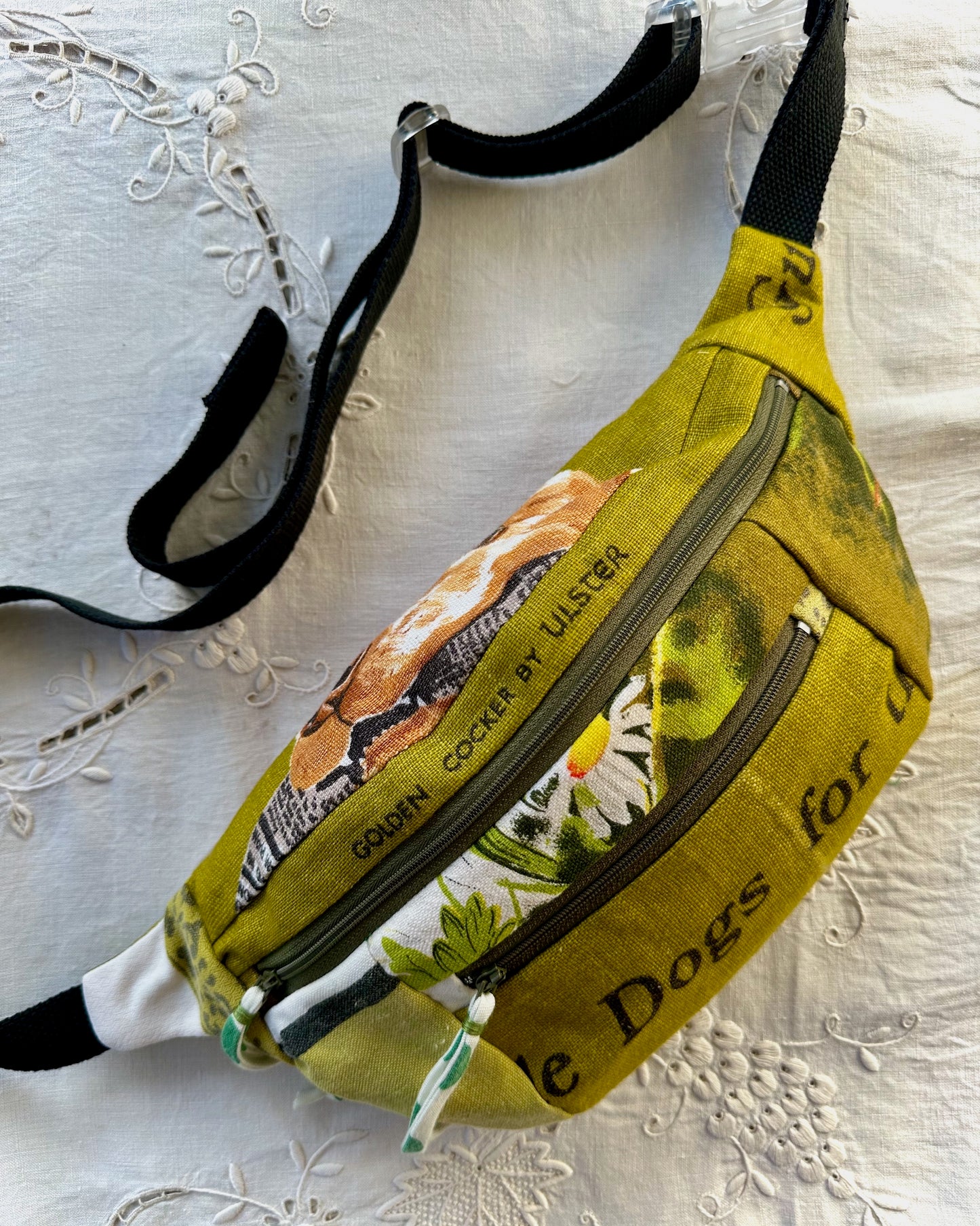 A yellow-green fanny pack featuring a printed graphic of a golden retriever, white daisies, and the text 'GOLDEN COCKER BY ULSTER' and 'e Dogs for u'. It has black straps and is laid flat on a white embroidered fabric background.