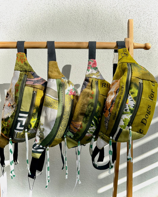 Four fashion bum bags with olive green and multicolor vintage print designs hang from a wooden display stand against a textured white wall.