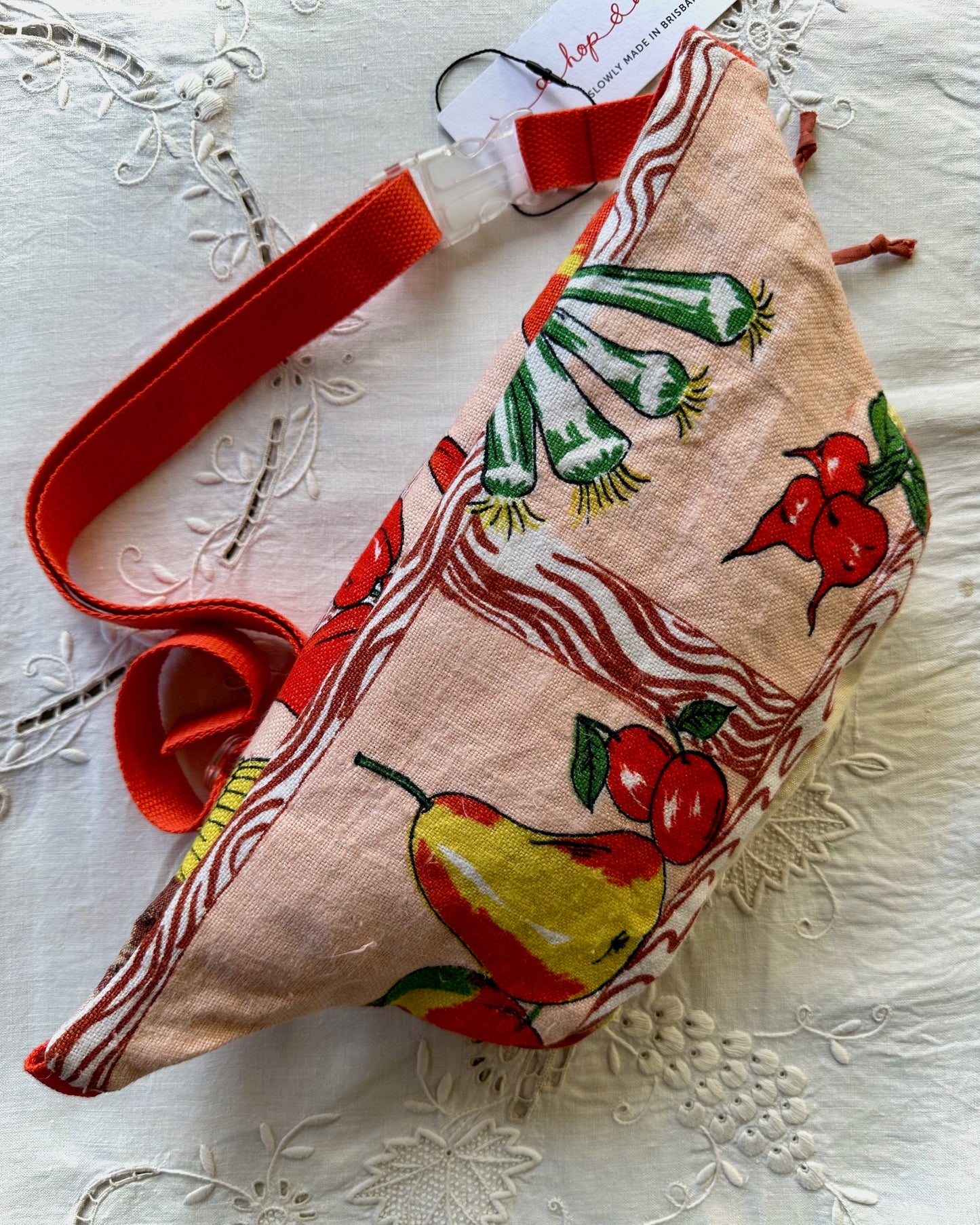 A small fabric pouch with a red strap, featuring a vintage-style print of pears, radishes, and green onions on a beige linen-like background, displayed on a white embroidered fabric surface.