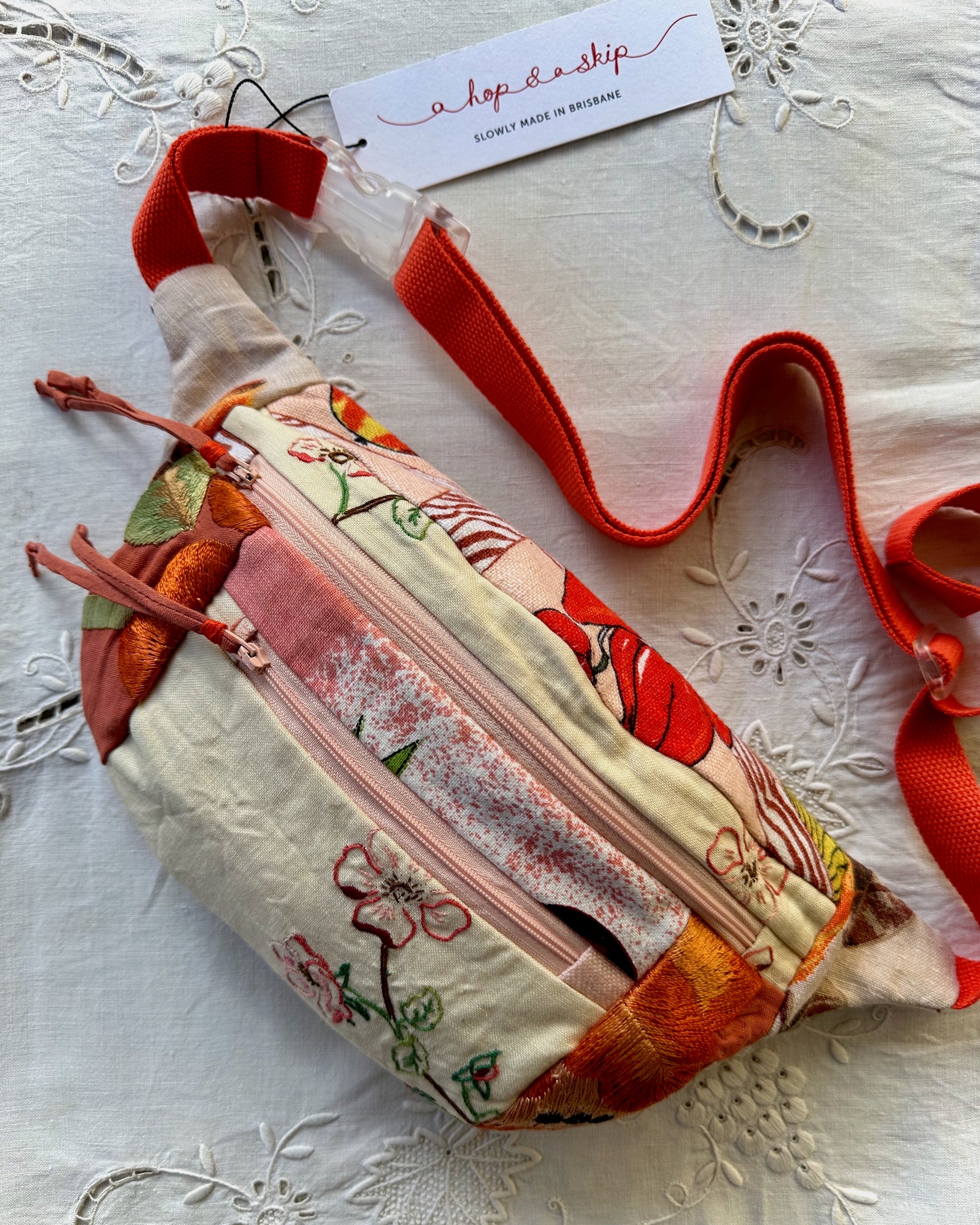 A handmade fabric crossbody bag with a colorful floral print, featuring a bright red-orange strap, a zippered front pocket, and a paper tag reading 'a hop and a skip. The bag is laid flat on a white embroidered lace fabric background.
