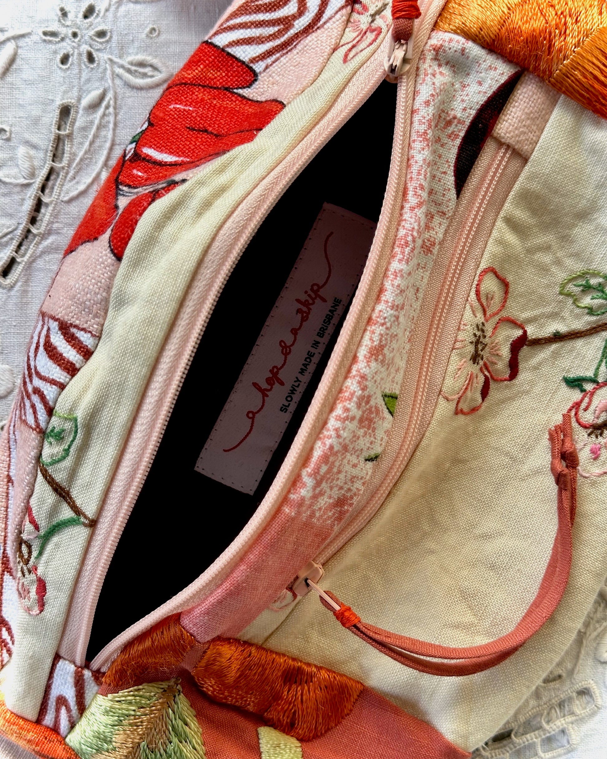 Close-up of a fabric zippered pouch with a cream base, pink floral embroidery, red and orange embroidered accents, and a visible interior label reading 'a hop and a skip'.
