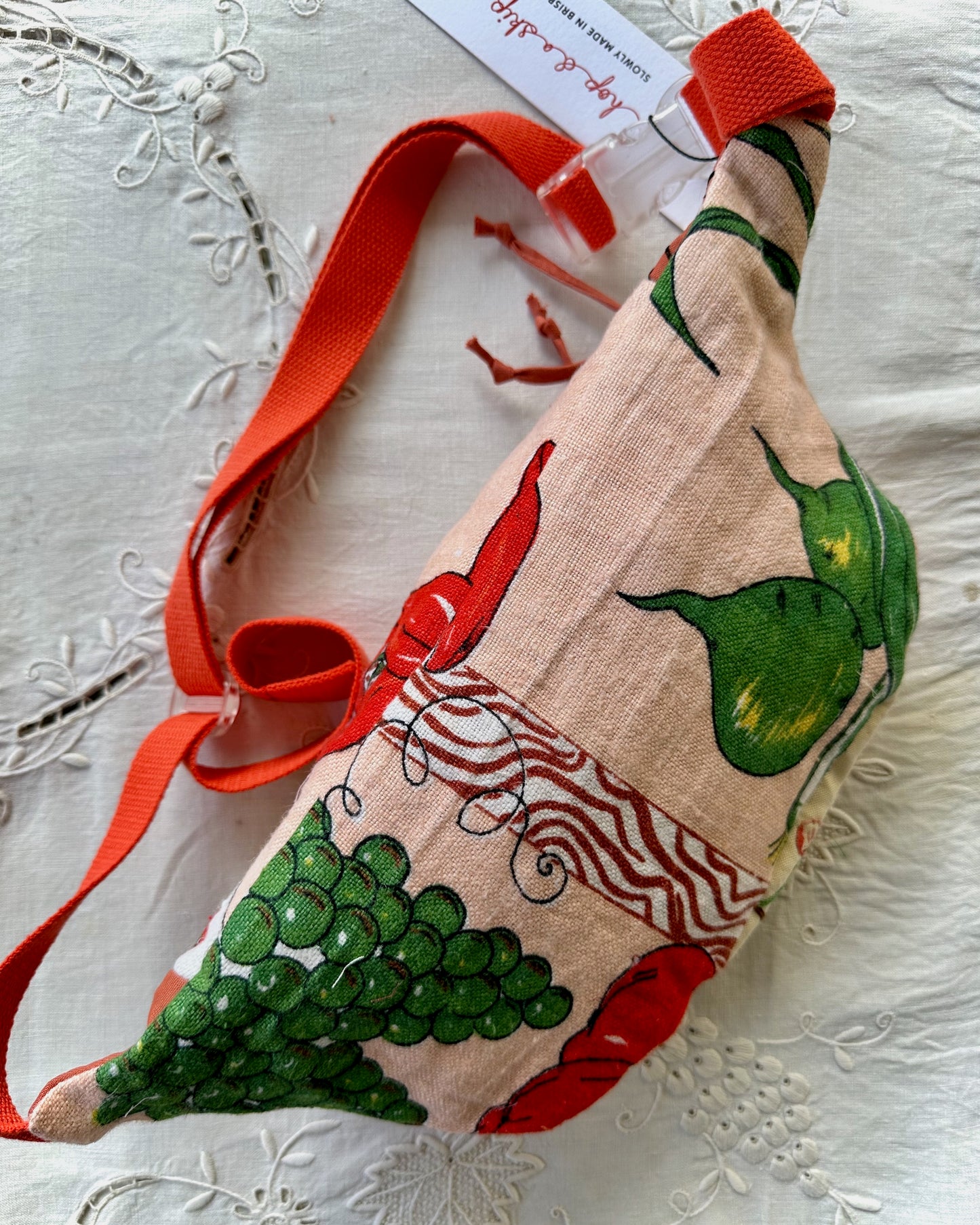 A handmade fabric bag with a red shoulder strap, featuring a printed pattern of red chili peppers, green grapes, and green pears on a beige linen background. The bag is displayed on a white embroidered fabric surface with its paper tag still attached.