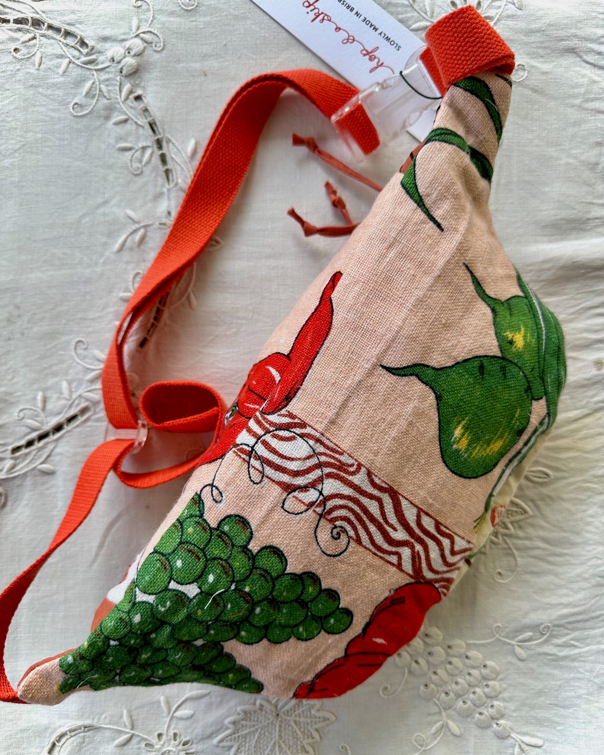 A handmade fabric bag with a red shoulder strap, featuring a printed pattern of red chili peppers, green grapes, and green pears on a beige linen background. The bag is displayed on a white embroidered fabric surface with its paper tag still attached.