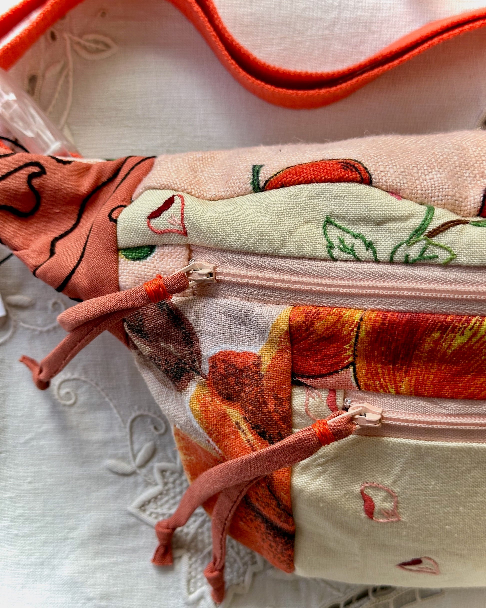 A close-up of a fabric pouch with a multicolor floral and fruit print, featuring orange zipper pulls and a peach-colored strap, resting on a white lace surface.