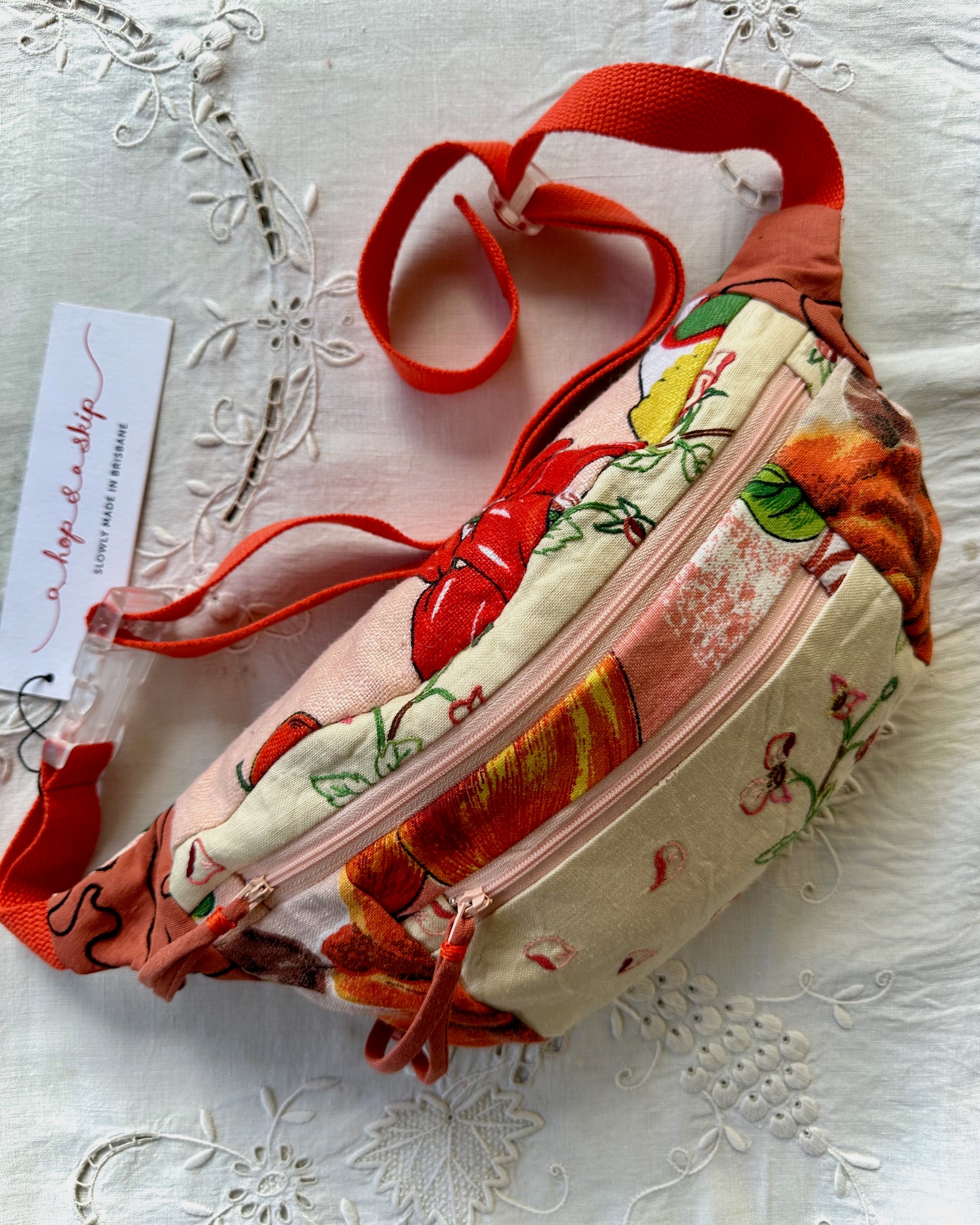 A fabric fanny pack with a colorful floral and fruit print, featuring a prominent red-orange strap and a pink zipper, displayed on a white embroidered lace background with a brand tag.