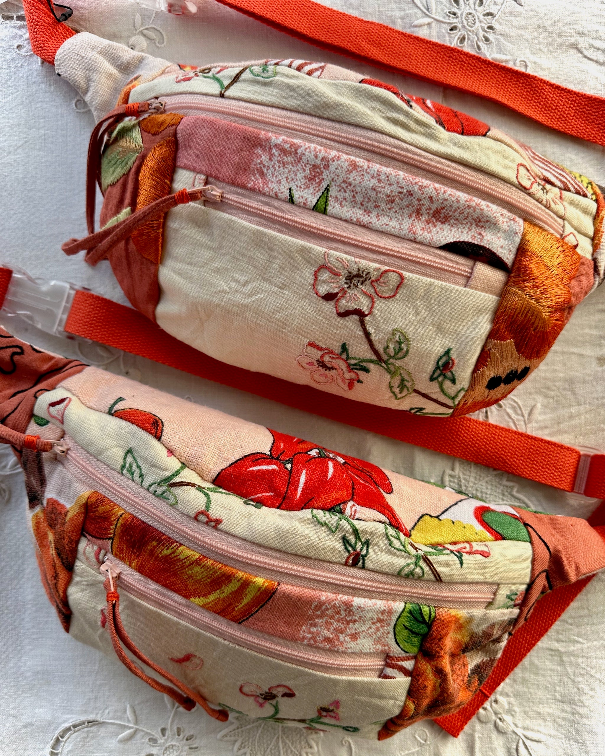 Two quilted fabric fanny packs with floral and fruit prints, featuring orange-red webbing straps, displayed on a white lace fabric background.