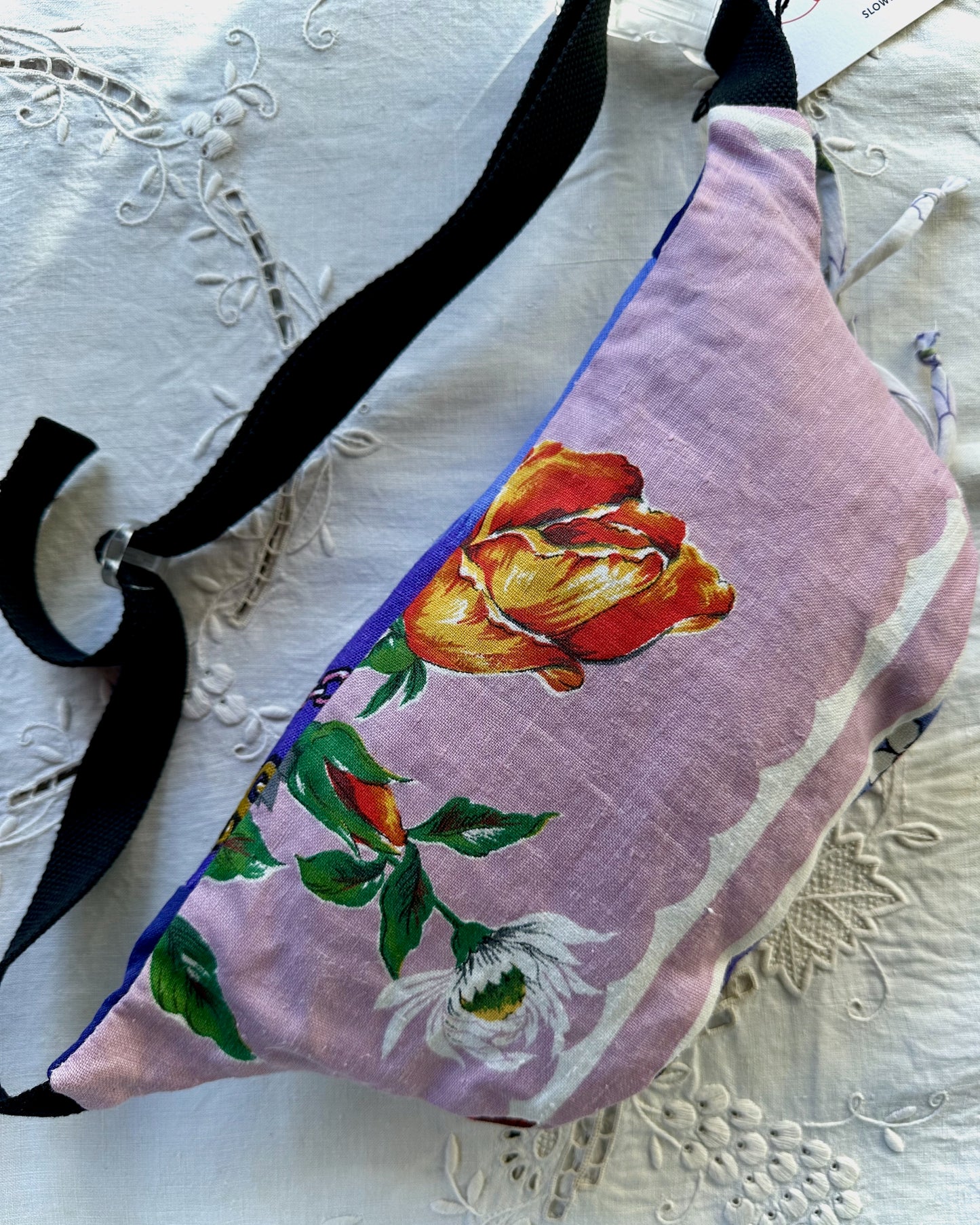 A fabric crossbody bag with a lavender-purple background, featuring a large orange and yellow rose, green leaves, and a white flower printed on the front. The bag has black straps and is laid flat on a white embroidered lace fabric.