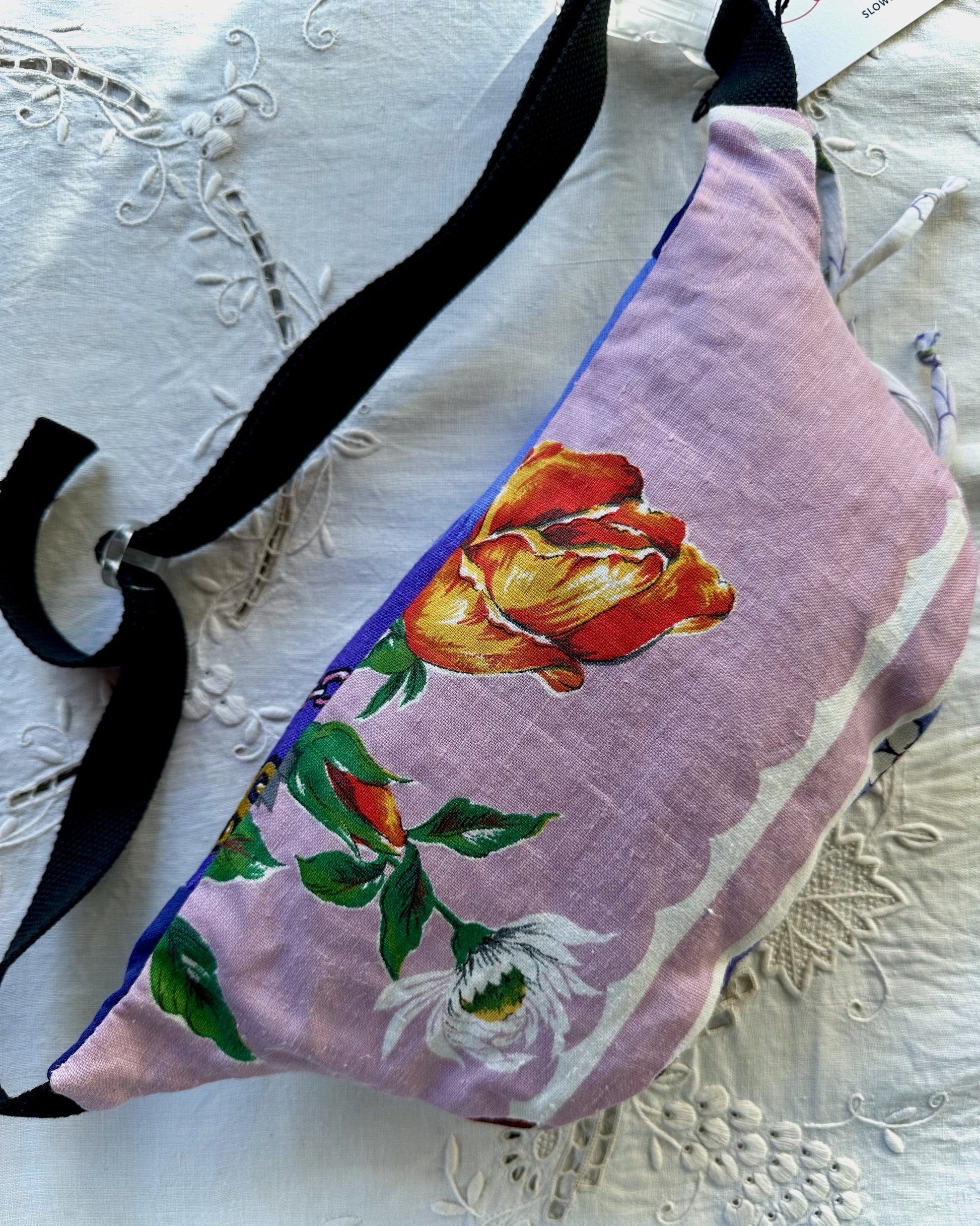 A fabric crossbody bag with a lavender-purple background, featuring a large orange and yellow rose, green leaves, and a white flower printed on the front. The bag has black straps and is laid flat on a white embroidered lace fabric.