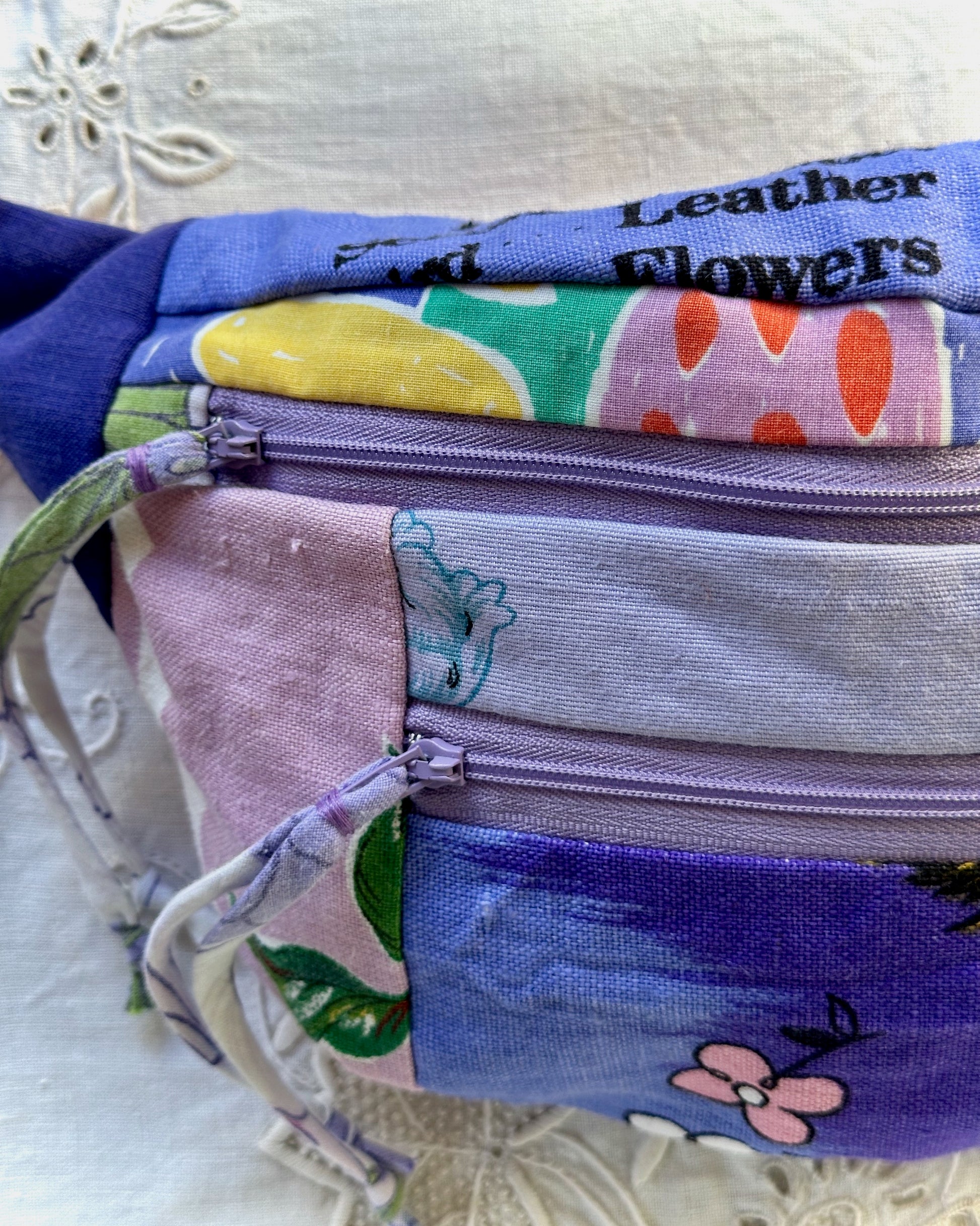 A close-up of a patchwork fabric pouch with multiple zippered compartments. The pouch features various colorful printed fabric panels in shades of purple, blue, pink, green, and yellow, with one panel showing the text 'Leather Flowers'.