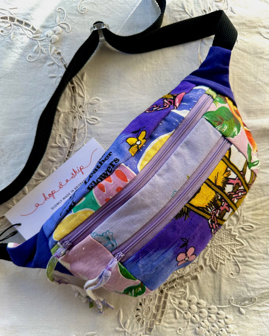 A colorful patchwork fanny pack with a black adjustable strap, featuring a purple background and a multicolor print of flowers, butterflies, and fruit. A white paper tag with the brand name 'a hop and a skip' is attached to the front.
