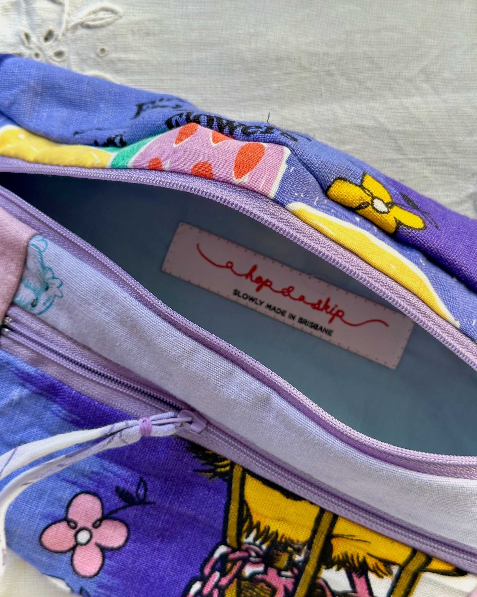 A close-up of a zippered fabric bumbag with a colorful patchwork print in purple, yellow, pink, and blue. The interior lining is light purple, and a sewn-on label reads 'a hop a skip' with 'slowly made in brisbane' underneath.