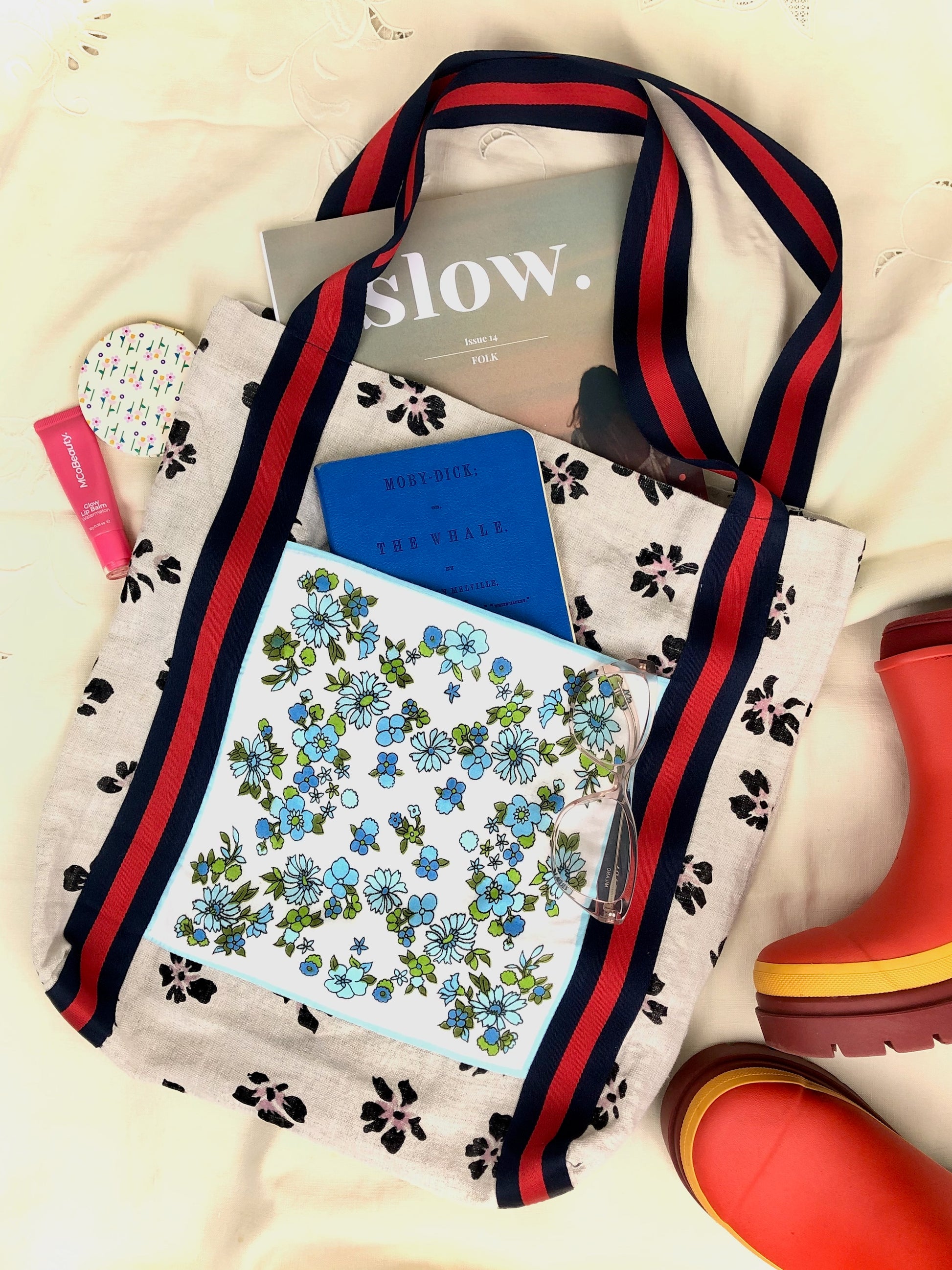 Flat lay of a handmade tote bag with Slow magazine, journal, lip gloss, reading glasses and Merry People gum boots