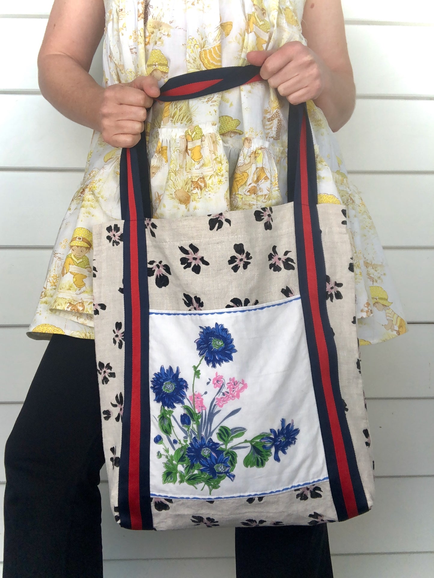 Woman wearing a yellow Sarah Kay babydoll dress and holding a floral tote bag with a large blue and pink floral design and read and navy striped straps